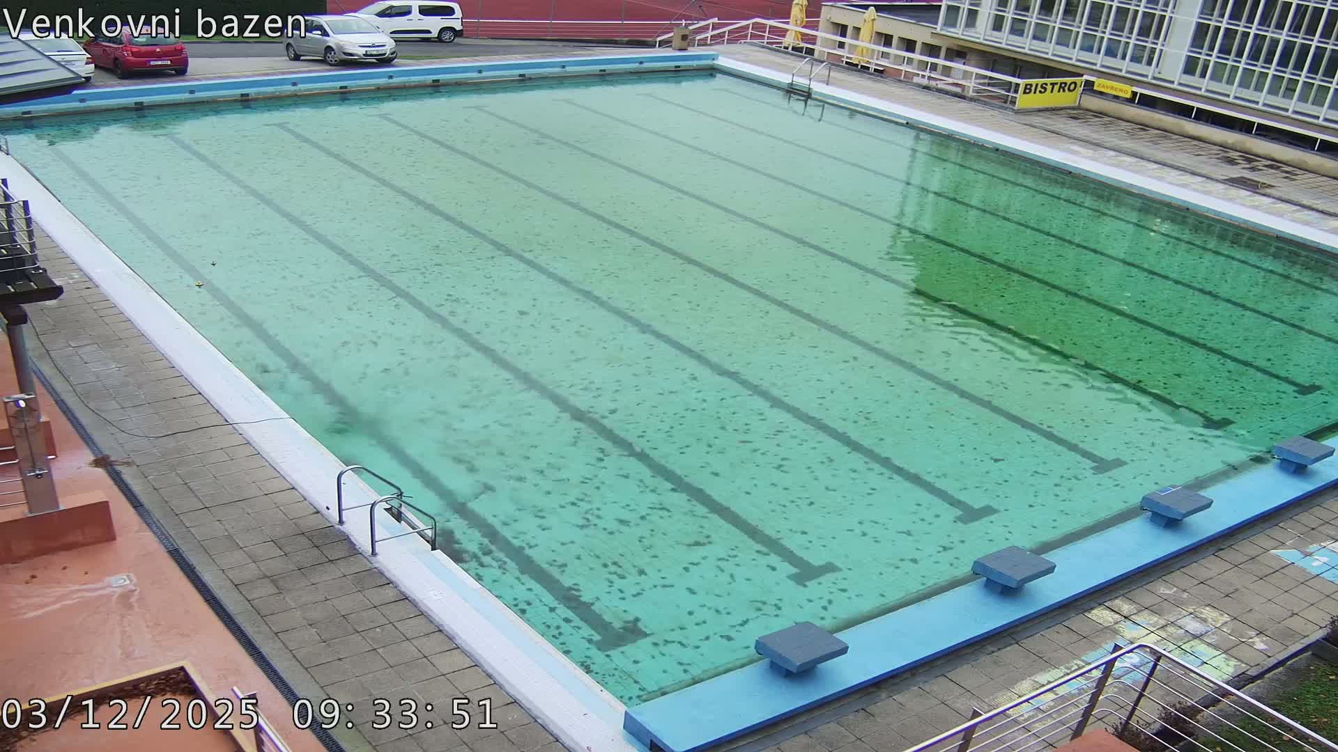 An outdoor swimming pool with visible lane lines and starting blocks is filled with murky greenish-blue water showing ripples from light rain, all under an overcast sky, with a paved deck, a building, and parked cars in the background.