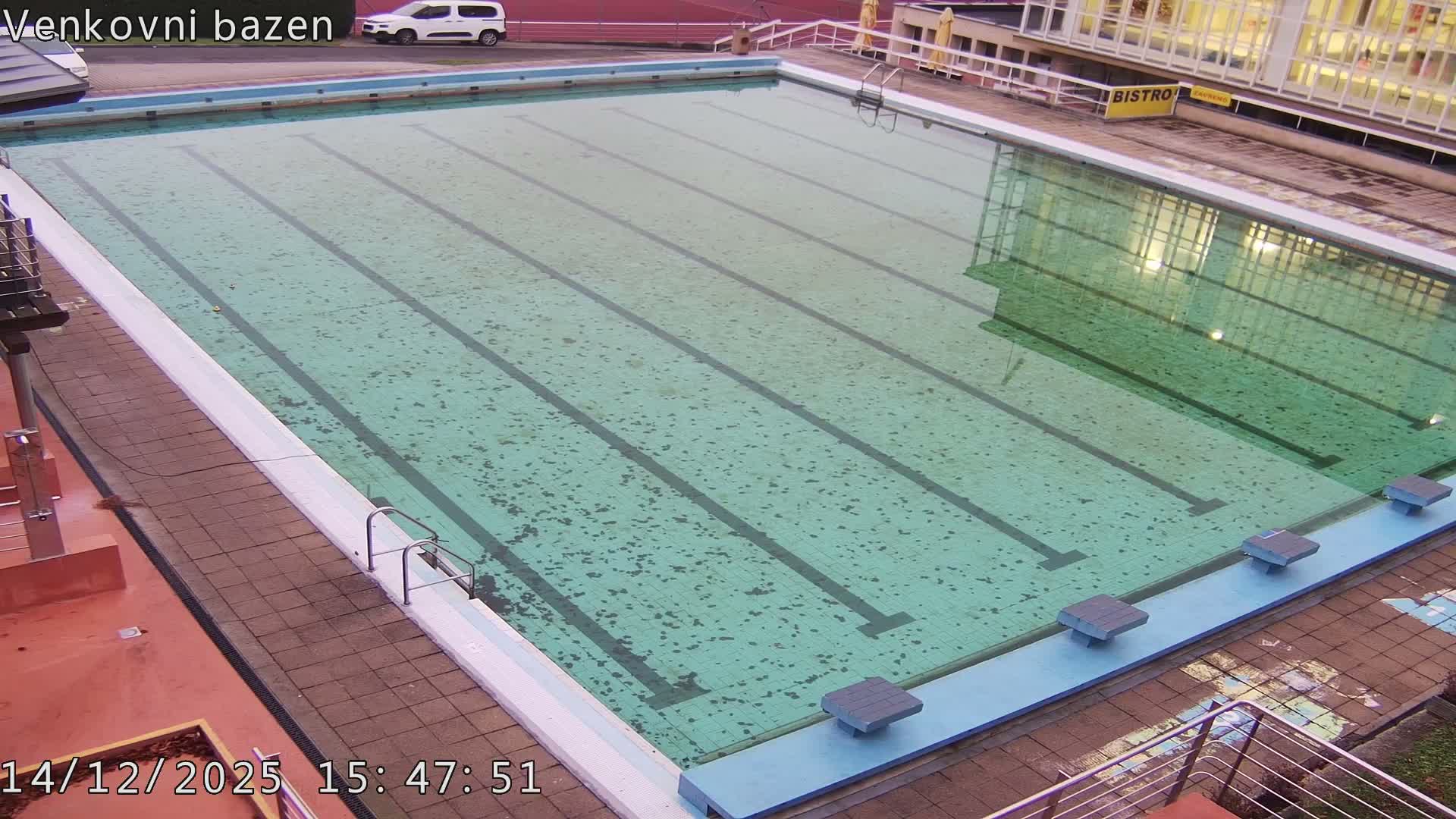 An outdoor swimming pool with visible lane lines and starting blocks is filled with murky greenish-blue water showing ripples from light rain, all under an overcast sky, with a paved deck, a building, and parked cars in the background.