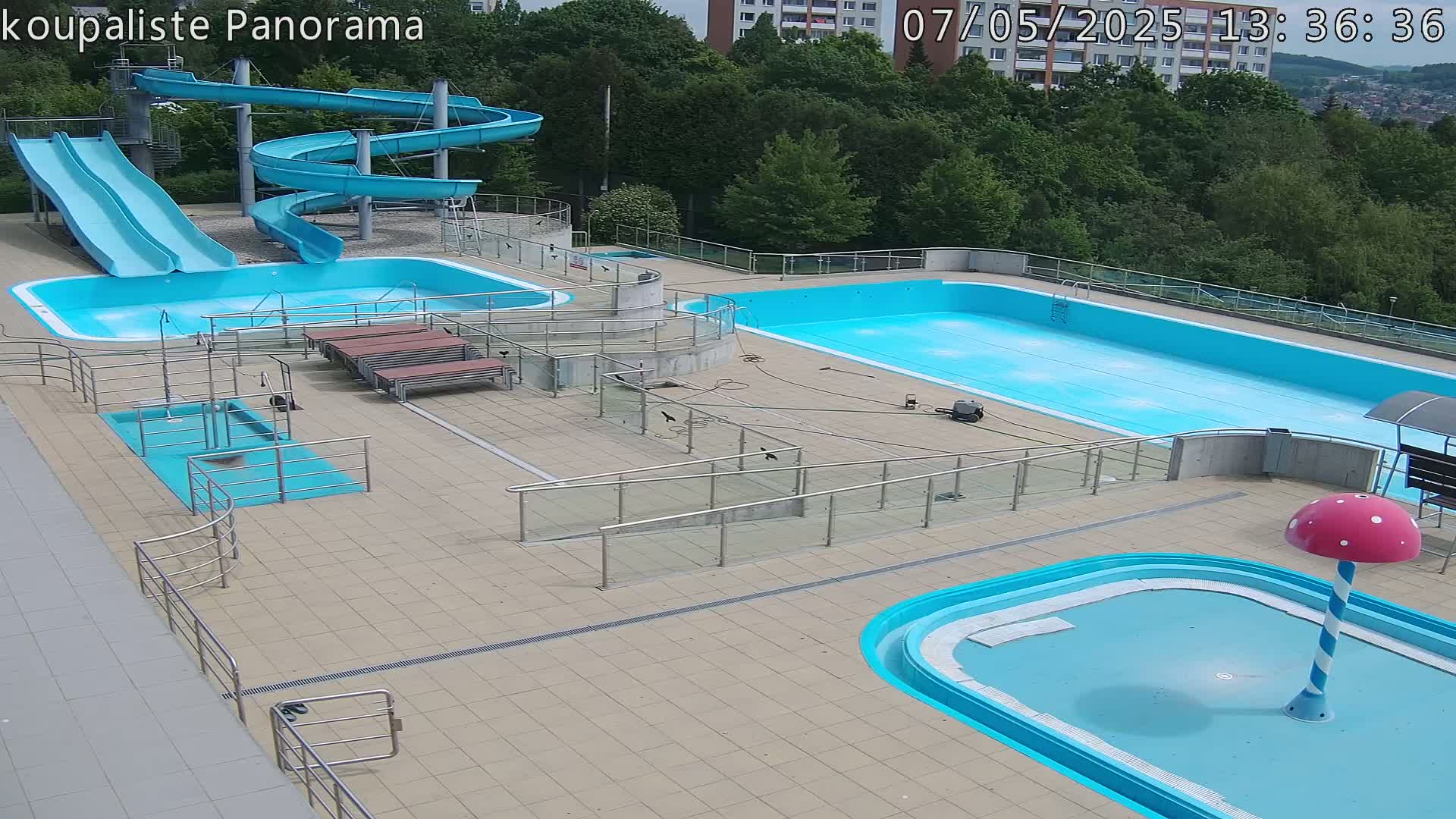 An outdoor pool complex with two large pools, two slides, a smaller wading pool, and several ramps and railings, on a sunny day.