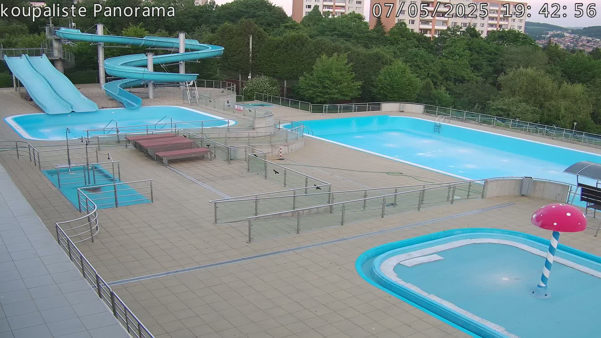 An outdoor pool complex with multiple pools, water slides, and a small children's pool, under a partly cloudy sky.