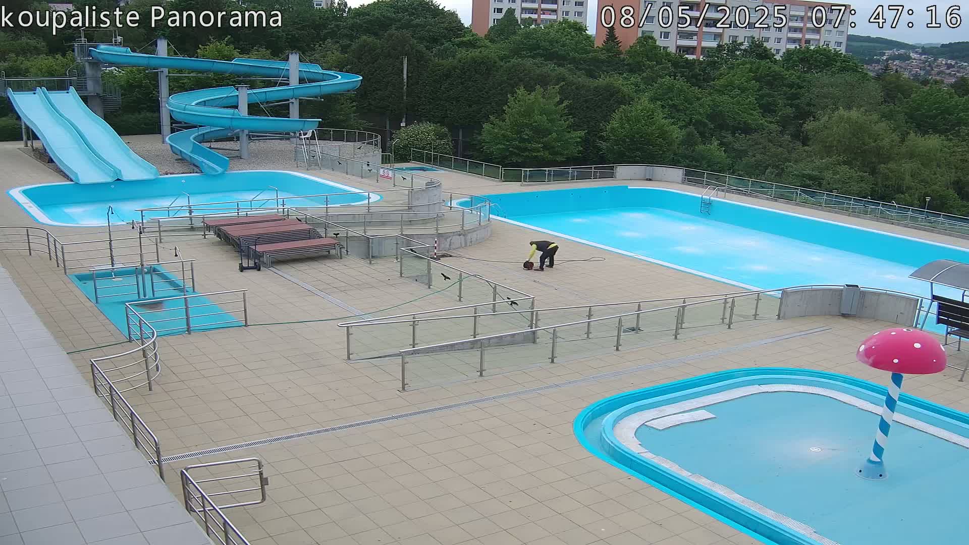 An outdoor pool complex with two large pools, several smaller pools, two slides, and a person working near one of the pools, on a mostly sunny day.