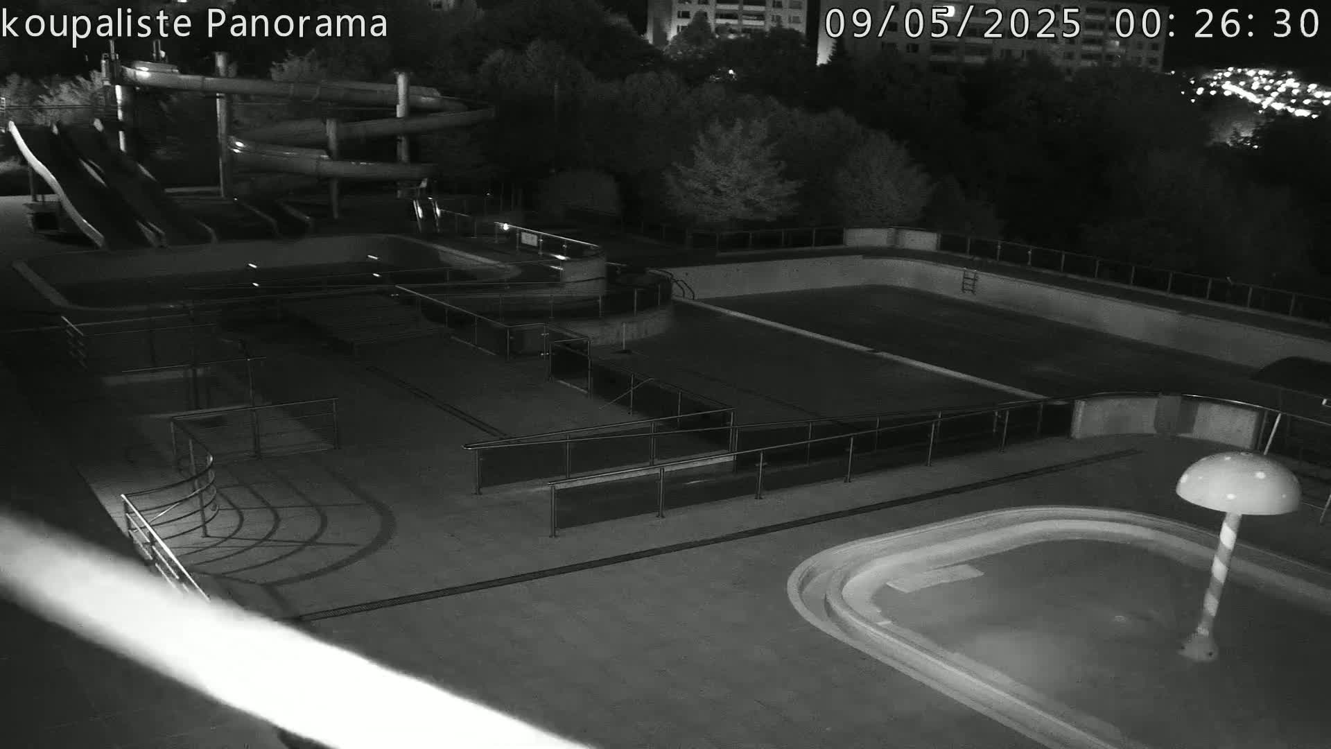 An outdoor nighttime view of an empty swimming complex with water slides and multiple pools, including a small pool with a mushroom-shaped fountain.
