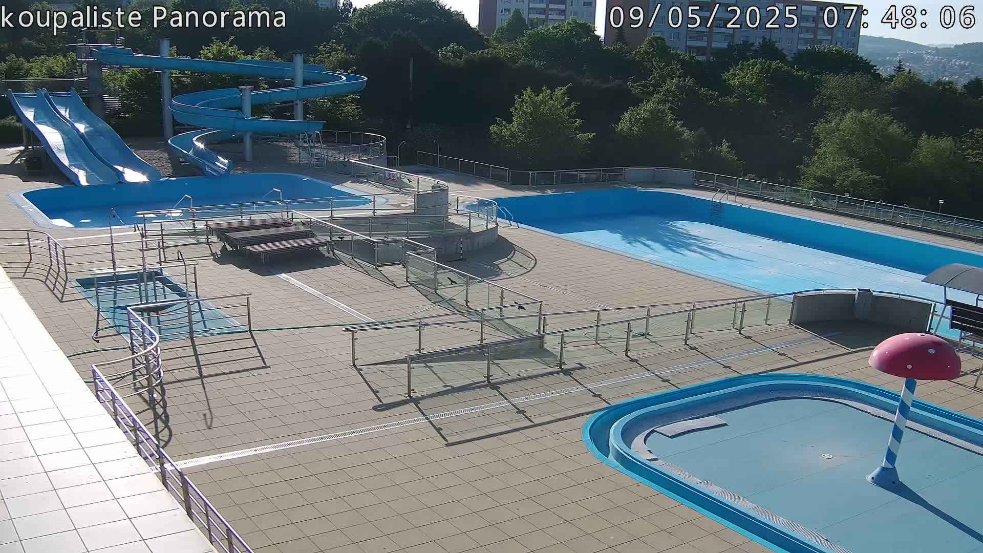 An outdoor swimming complex with multiple pools, slides, and a small mushroom-shaped water feature, bathed in sunlight.