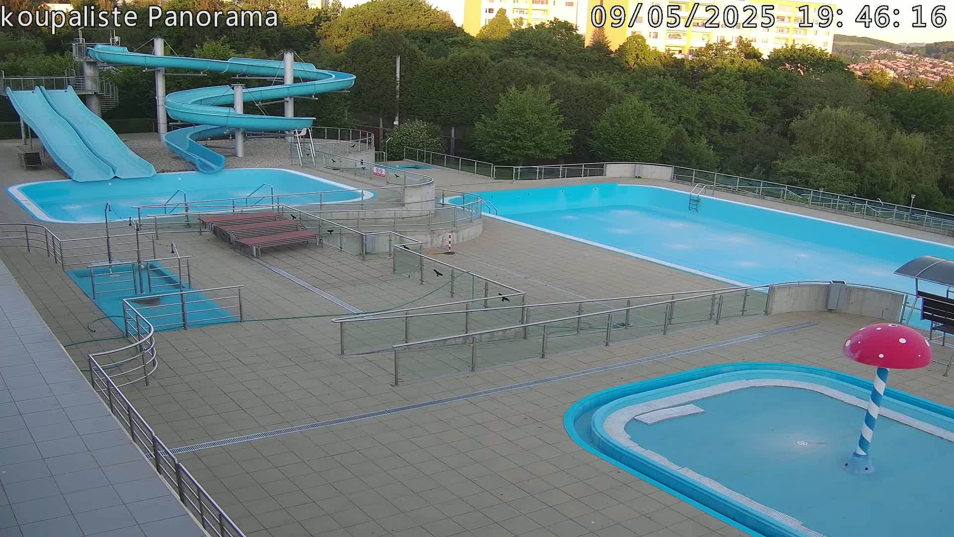 An outdoor swimming complex at dusk features several pools of varying sizes, two water slides, and a small mushroom-shaped water feature.