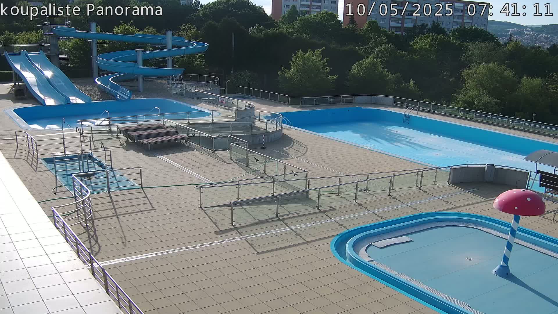 An outdoor pool complex with multiple pools, water slides, and accessibility features is shown under sunny conditions.