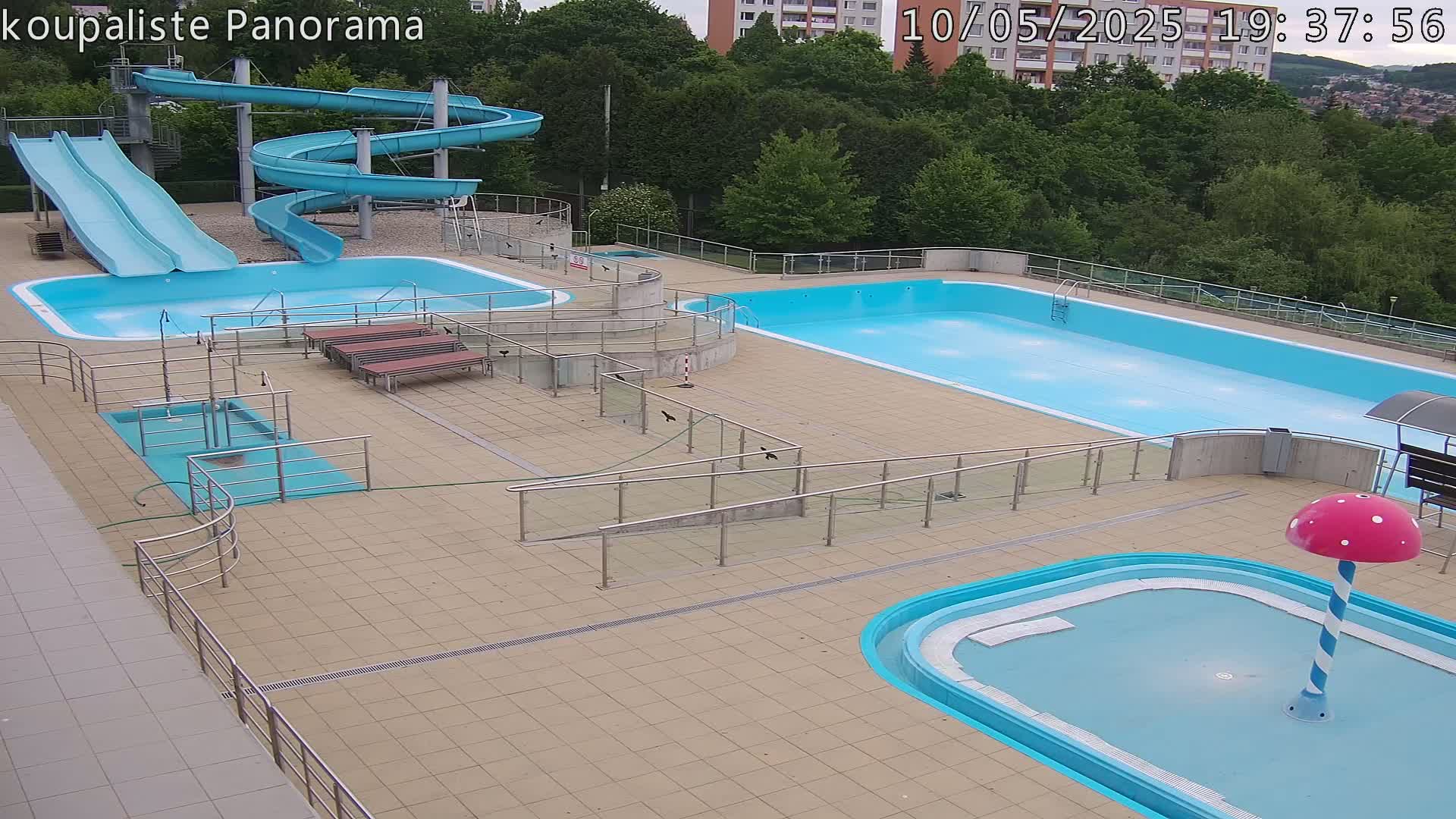 An outdoor pool complex with two large pools, two slides, a small wading pool with a mushroom-shaped fountain, and several benches, under a mostly sunny sky.