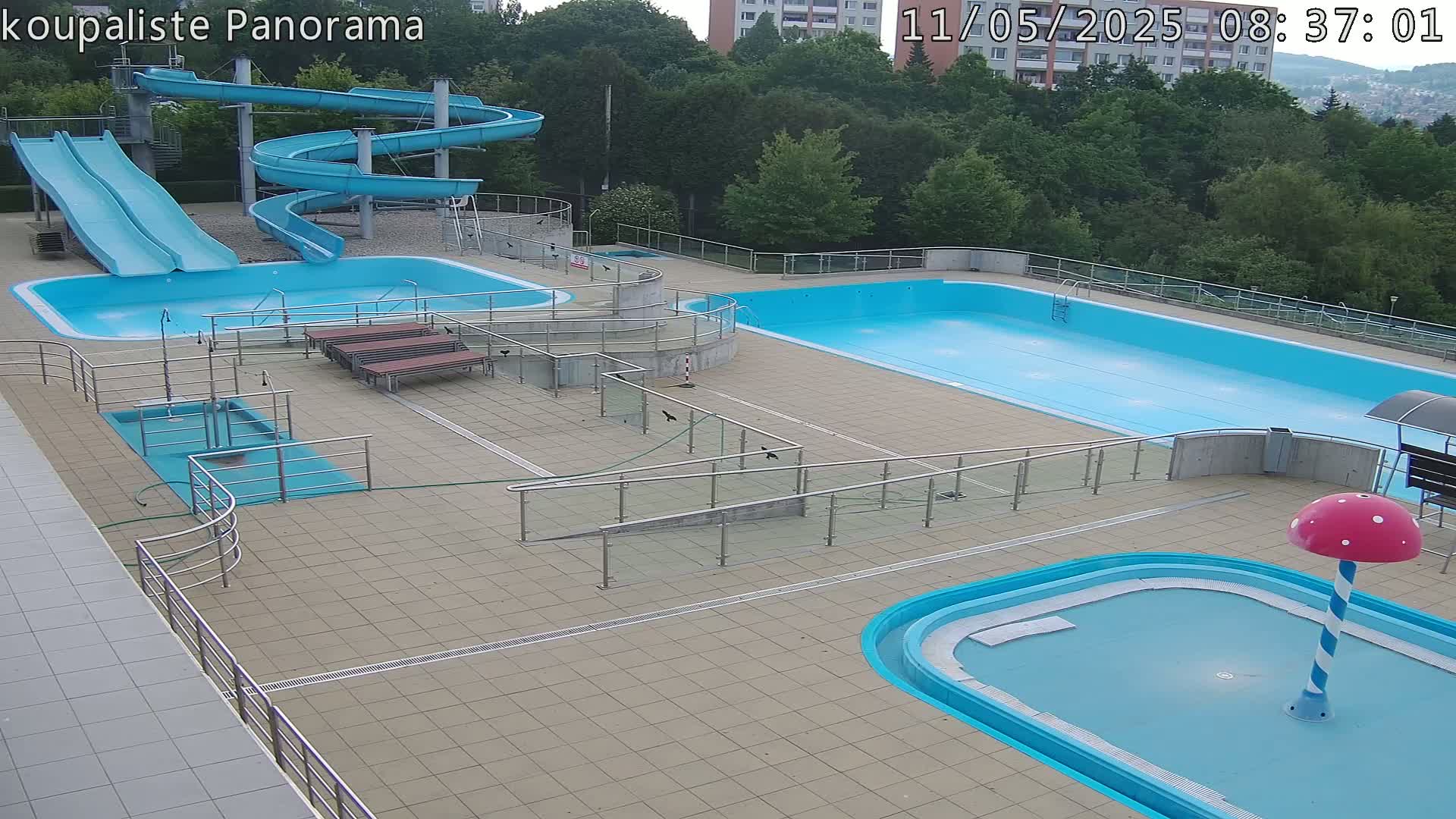 An outdoor pool complex with multiple pools, slides, and a small mushroom-shaped water feature, under a partly cloudy sky.