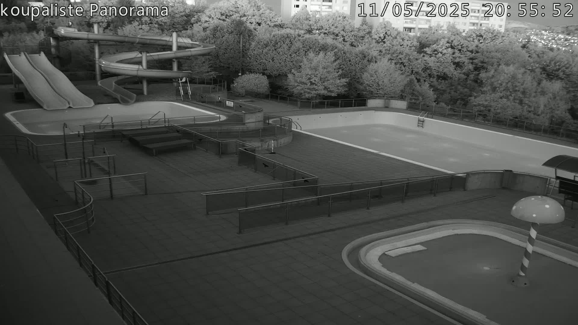 An outdoor pool complex at night, showing two large pools, several water slides, and a small wading pool, all surrounded by trees.