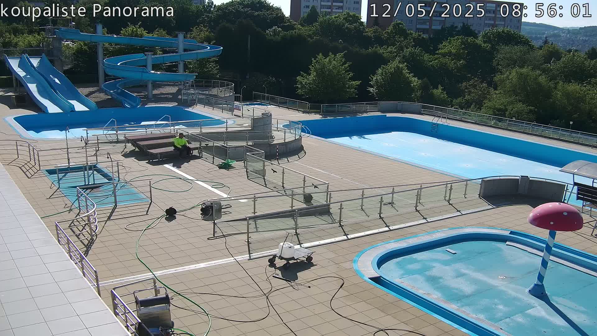 An outdoor swimming pool complex with multiple pools, water slides, and a person sitting on a bench, under a sunny sky.