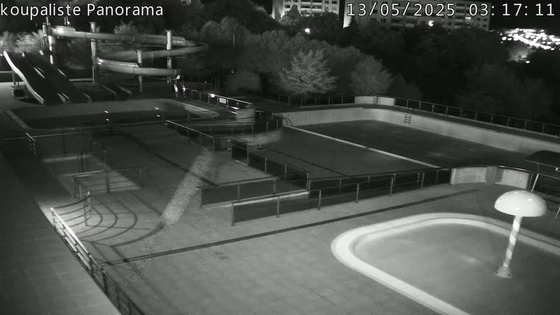 An empty outdoor swimming pool complex at night, featuring several pools, water slides, and railings, is illuminated by artificial light.