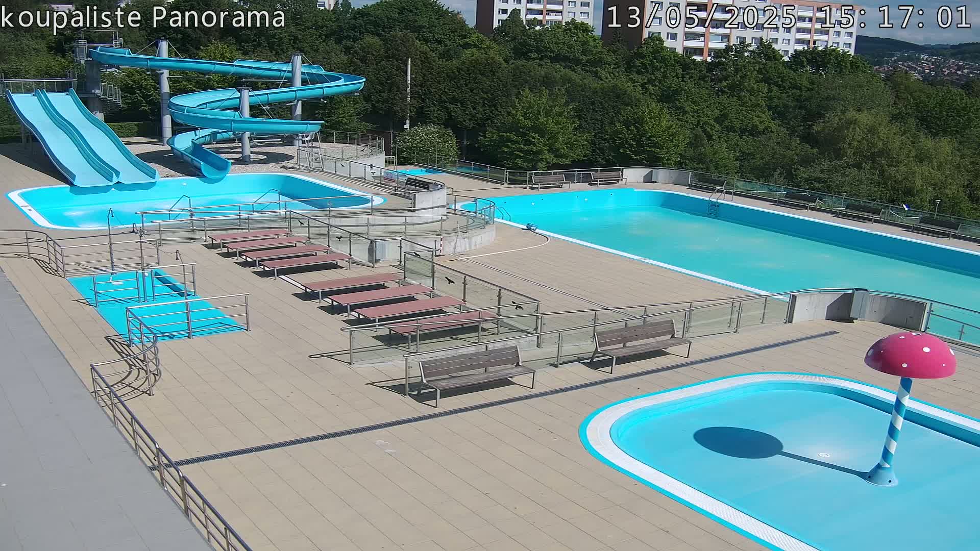 An outdoor pool complex on a sunny day features multiple pools of varying sizes, water slides, and several benches surrounding the pools.