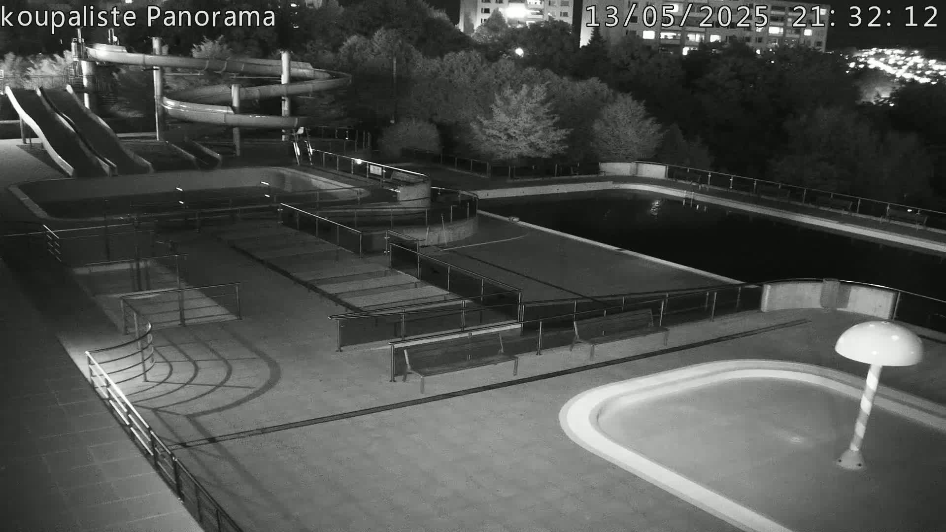 An outdoor swimming pool complex at night, featuring water slides, multiple pools, and benches, is illuminated by artificial light.