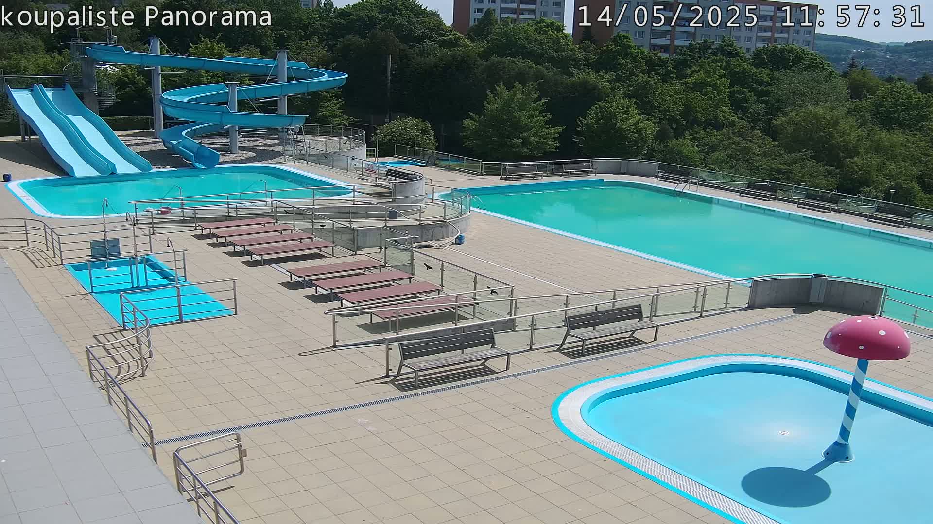 An outdoor pool complex with two large swimming pools, several smaller pools, water slides, and benches on a sunny day.