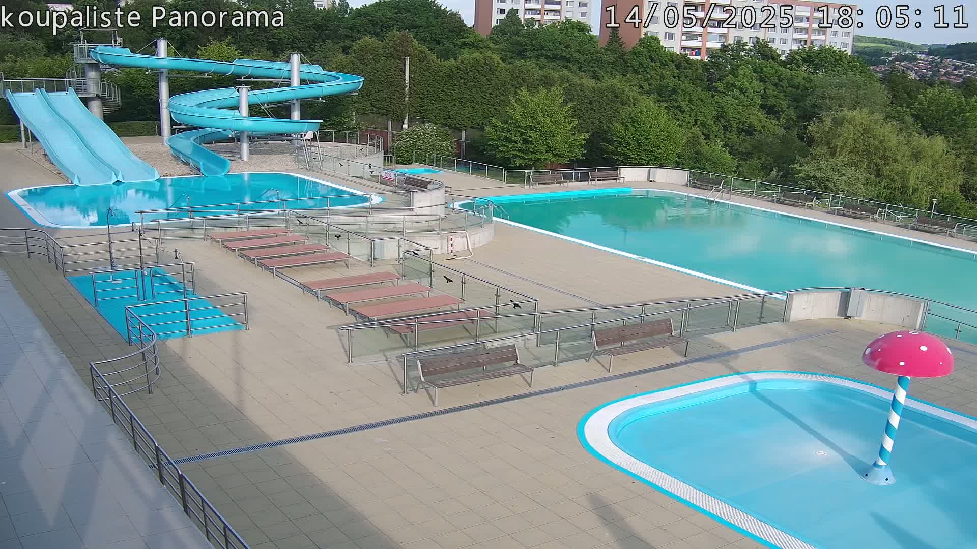An outdoor pool complex with two large pools, a smaller children's pool with a mushroom-shaped water feature, two slides, and several benches, all under sunny conditions.