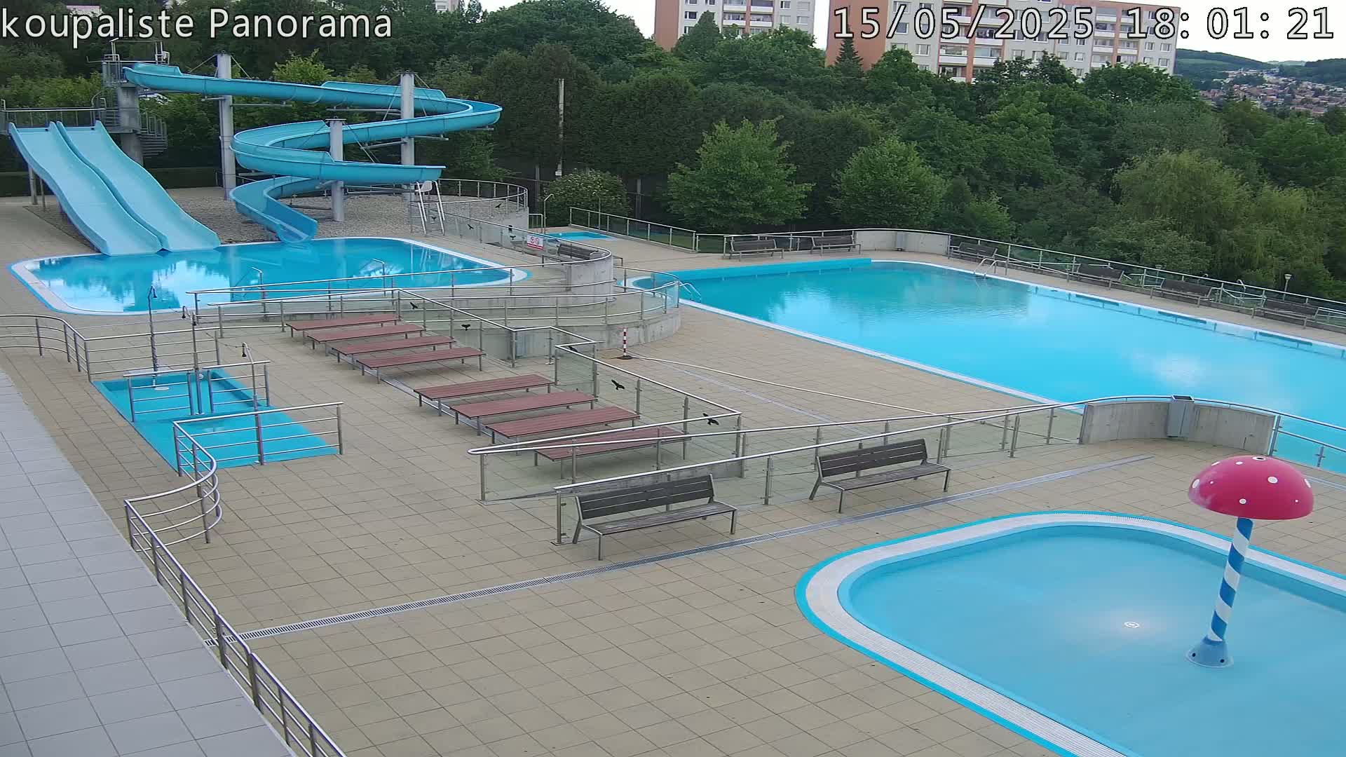 An outdoor pool complex with two large pools, several smaller pools, two slides, and several benches under a partly cloudy sky.