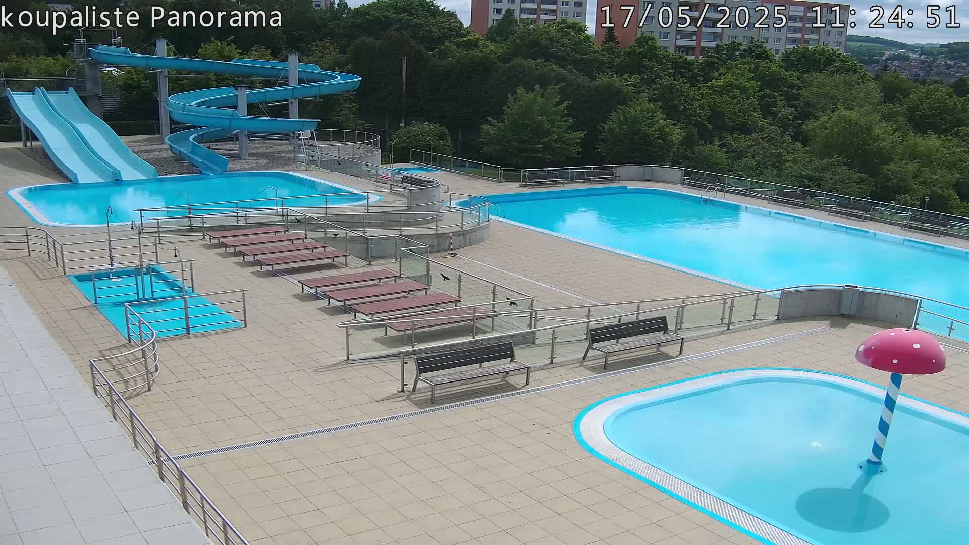 An outdoor pool complex with two large pools, a smaller wading pool with a mushroom-shaped fountain, two water slides, and several benches on a tiled deck.