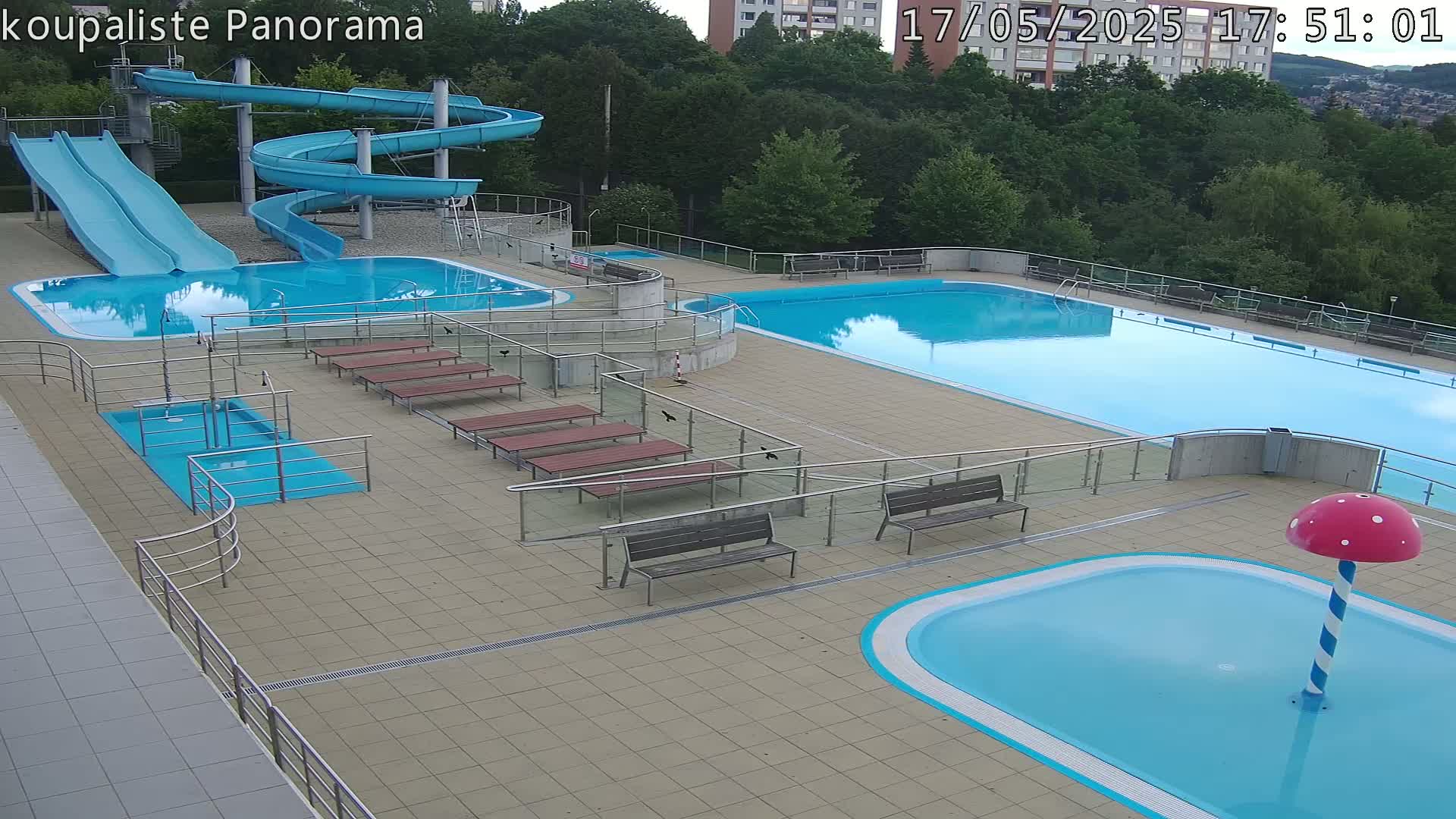 An outdoor swimming pool complex with two large pools, several smaller pools, water slides, and benches, under a clear sky.