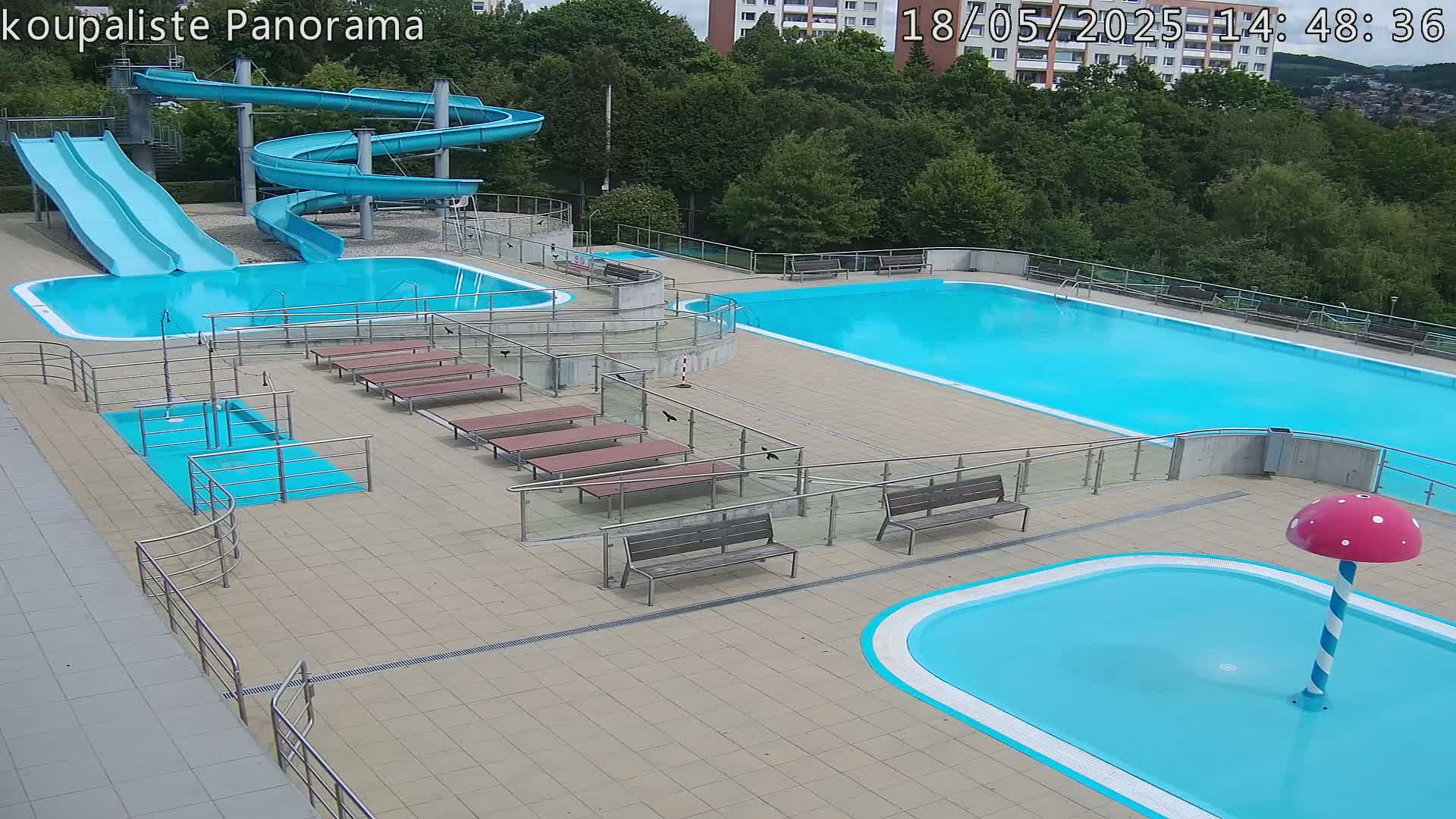 An outdoor pool complex with two large pools, two slides, several benches, and a small wading pool with a mushroom-shaped water feature on a sunny day.