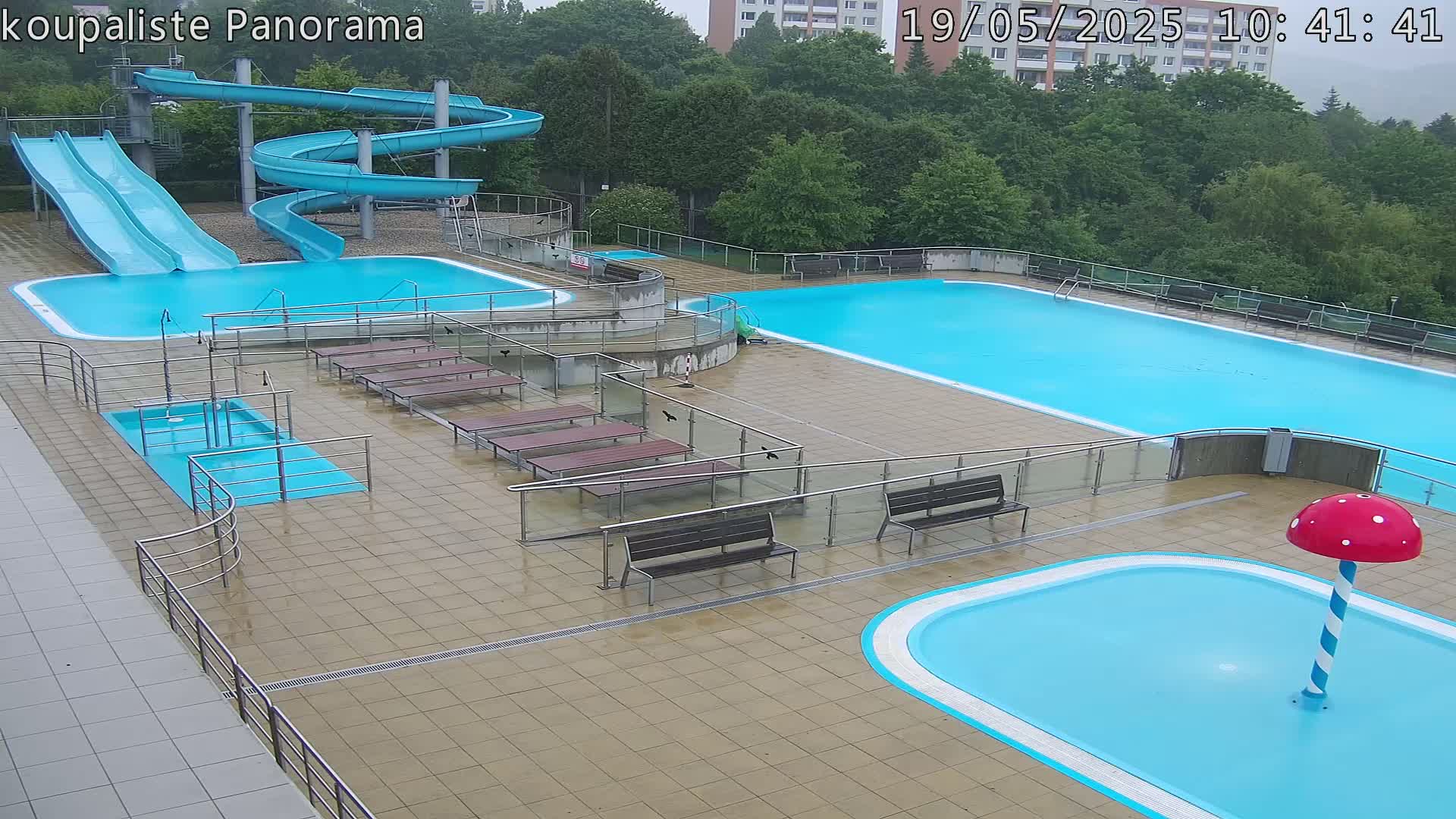 An outdoor pool complex with two large swimming pools, several smaller pools, water slides, and benches, all wet from recent rain.