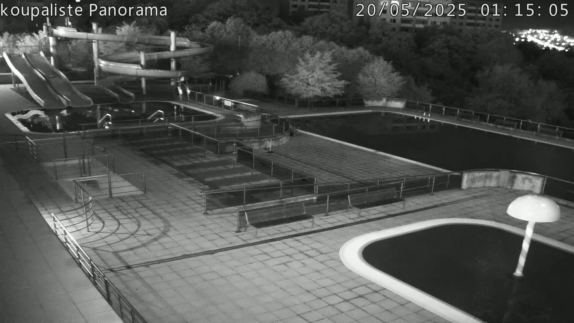 A nighttime, black-and-white view of an empty outdoor swimming complex with water slides and several pools, surrounded by trees.
