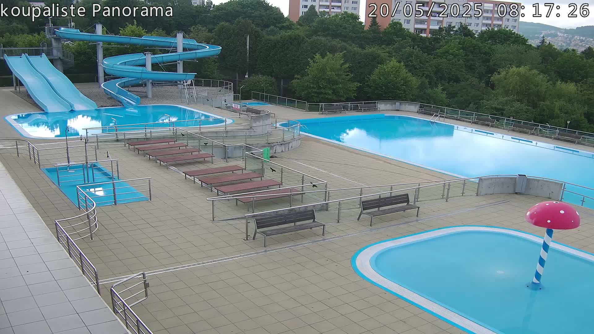 An outdoor pool complex with multiple pools, water slides, and benches, under partly cloudy skies.