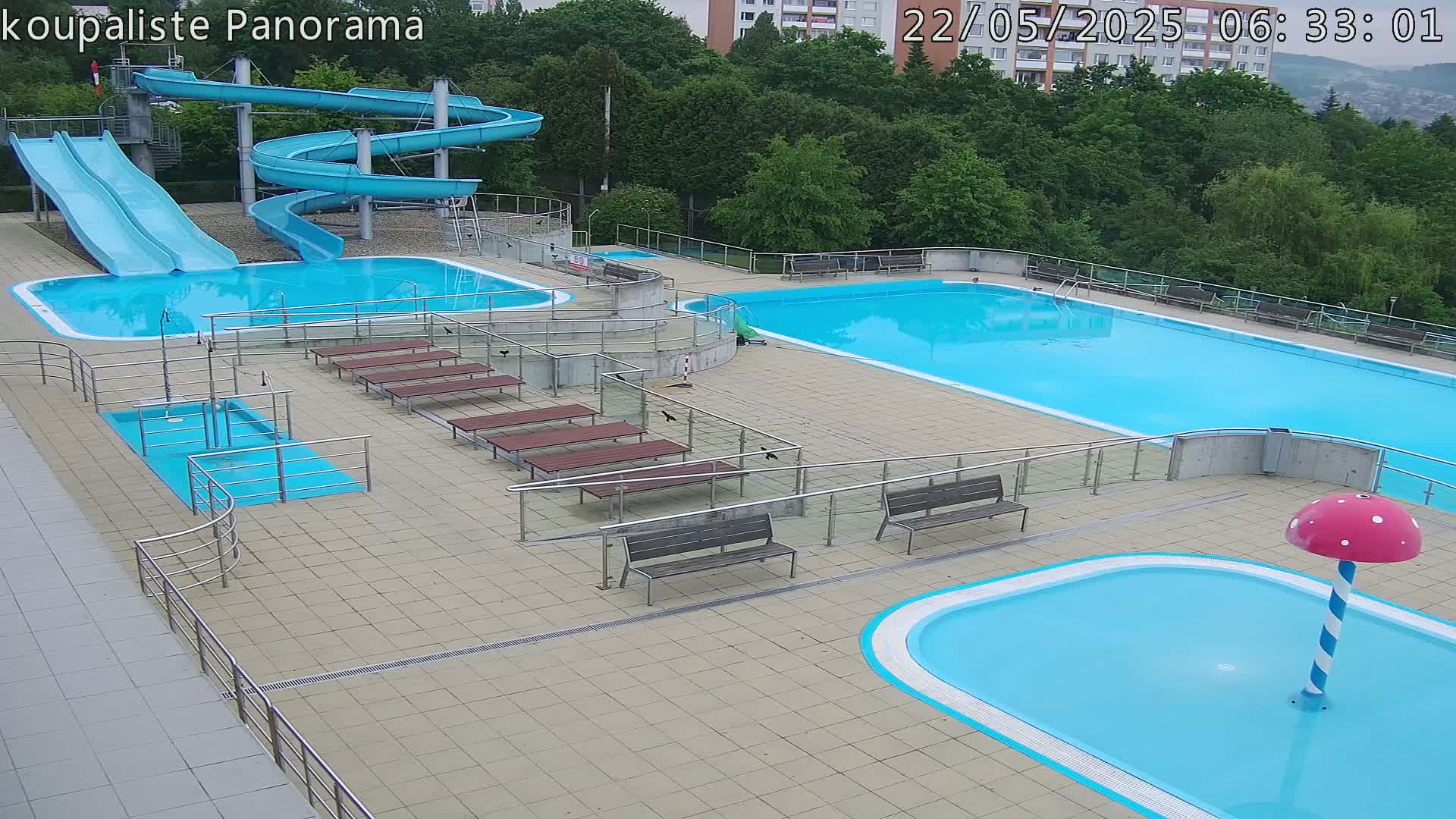 An outdoor pool complex with multiple pools, slides, and benches, under a partly cloudy sky.