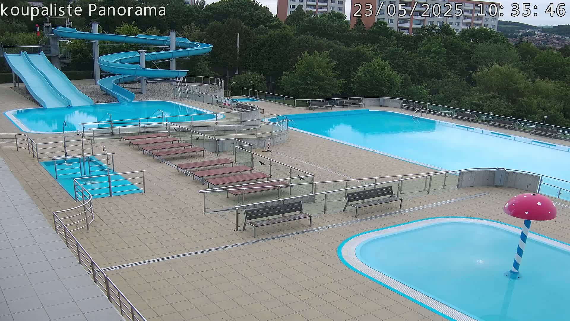 An outdoor swimming pool complex with two large pools, two water slides, a small wading pool with a mushroom-shaped fountain, and several benches under a mostly sunny sky.