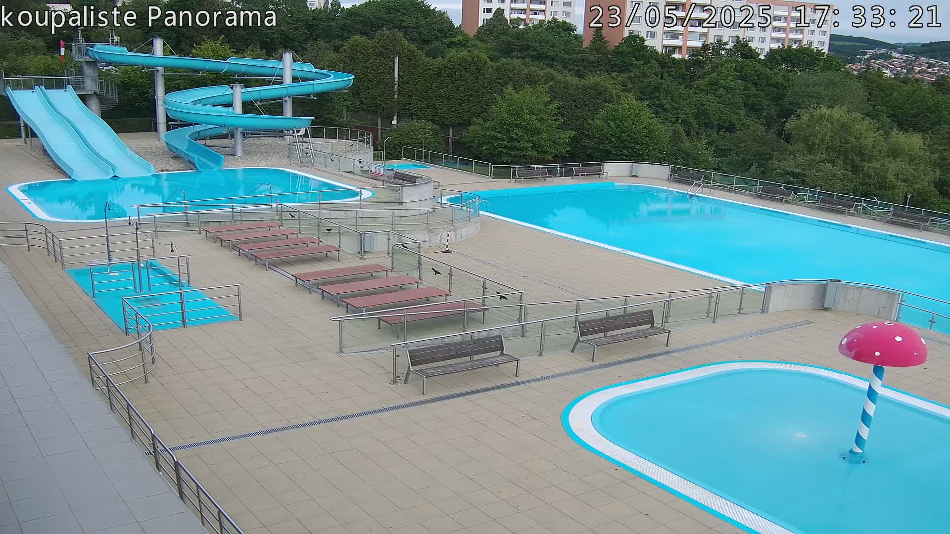 An outdoor pool complex with several pools of varying sizes, slides, benches, and a mushroom-shaped water feature, under a partly cloudy sky.