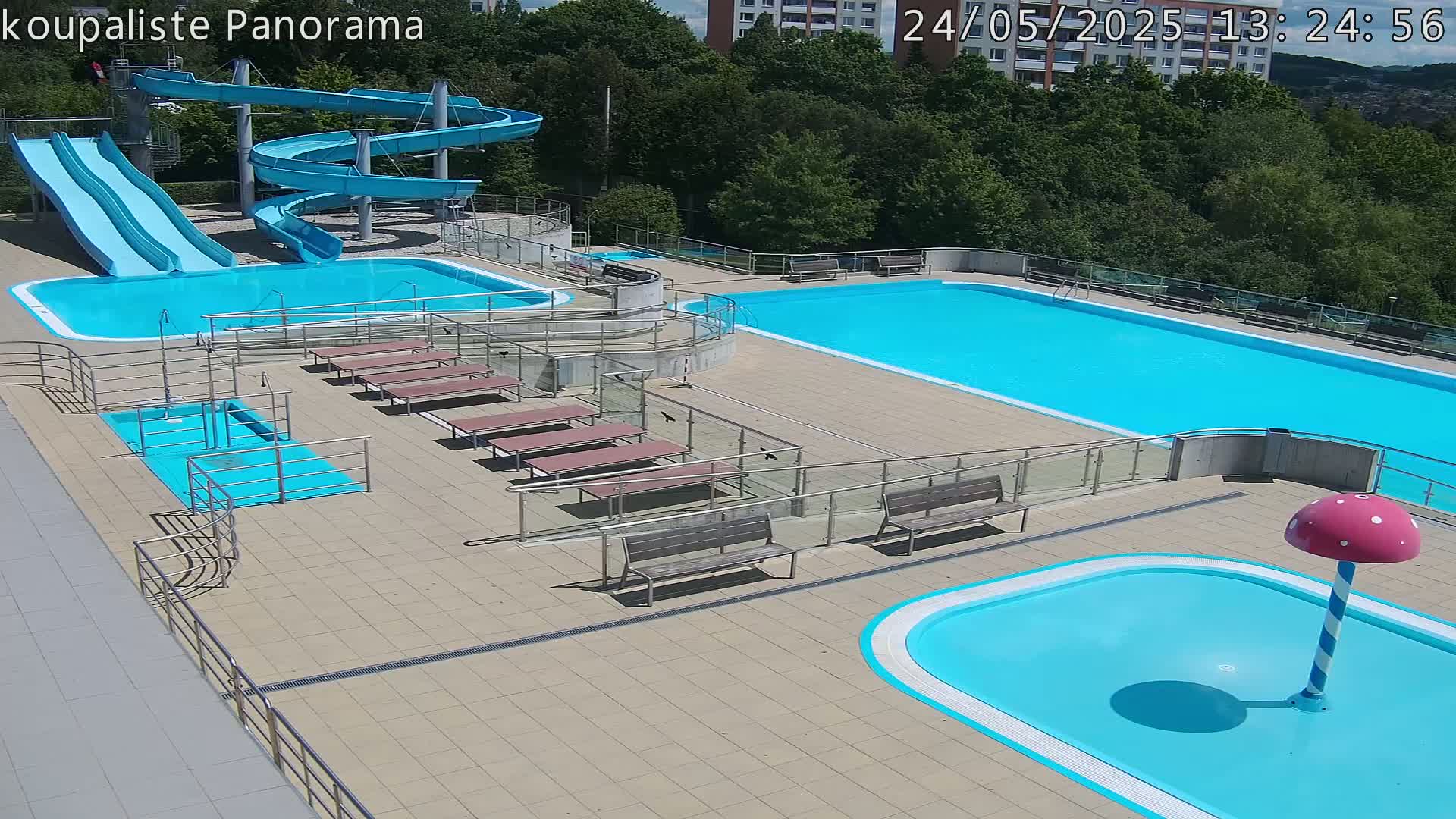 An outdoor pool complex with two large swimming pools, two slides, a small wading pool with a mushroom-shaped water feature, and several benches under a sunny sky.