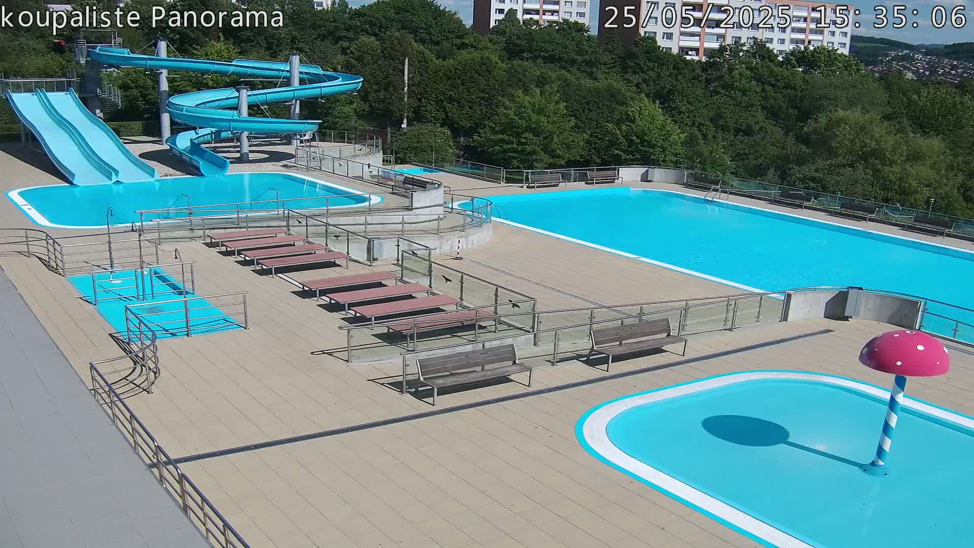 An outdoor pool complex on a sunny day features two large swimming pools, a smaller children's pool with a mushroom-shaped fountain, two slides, and several benches and railings.