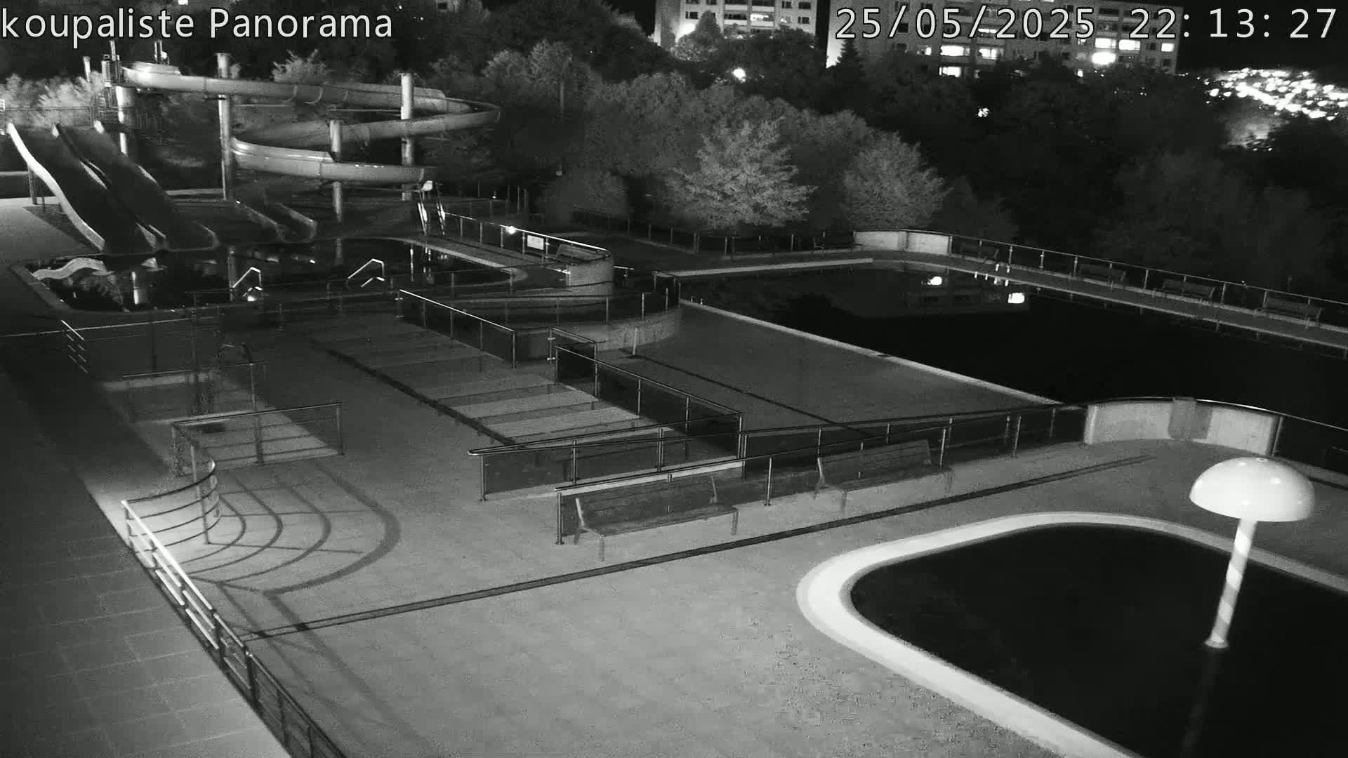 An empty outdoor swimming pool complex at night, featuring water slides and benches, is illuminated under artificial light.