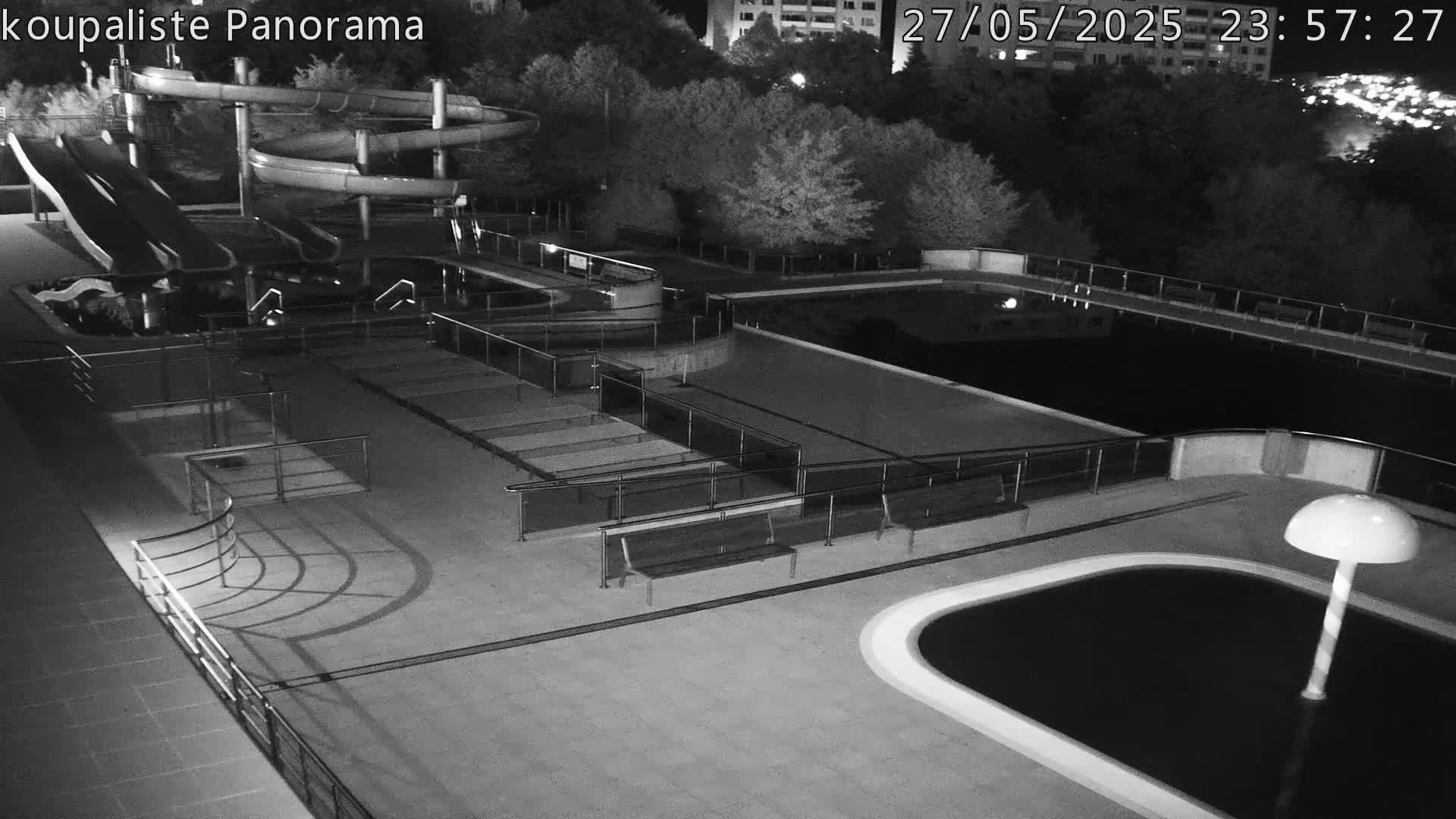 An outdoor water park at night, featuring several pools, water slides, and benches, is illuminated by artificial light.