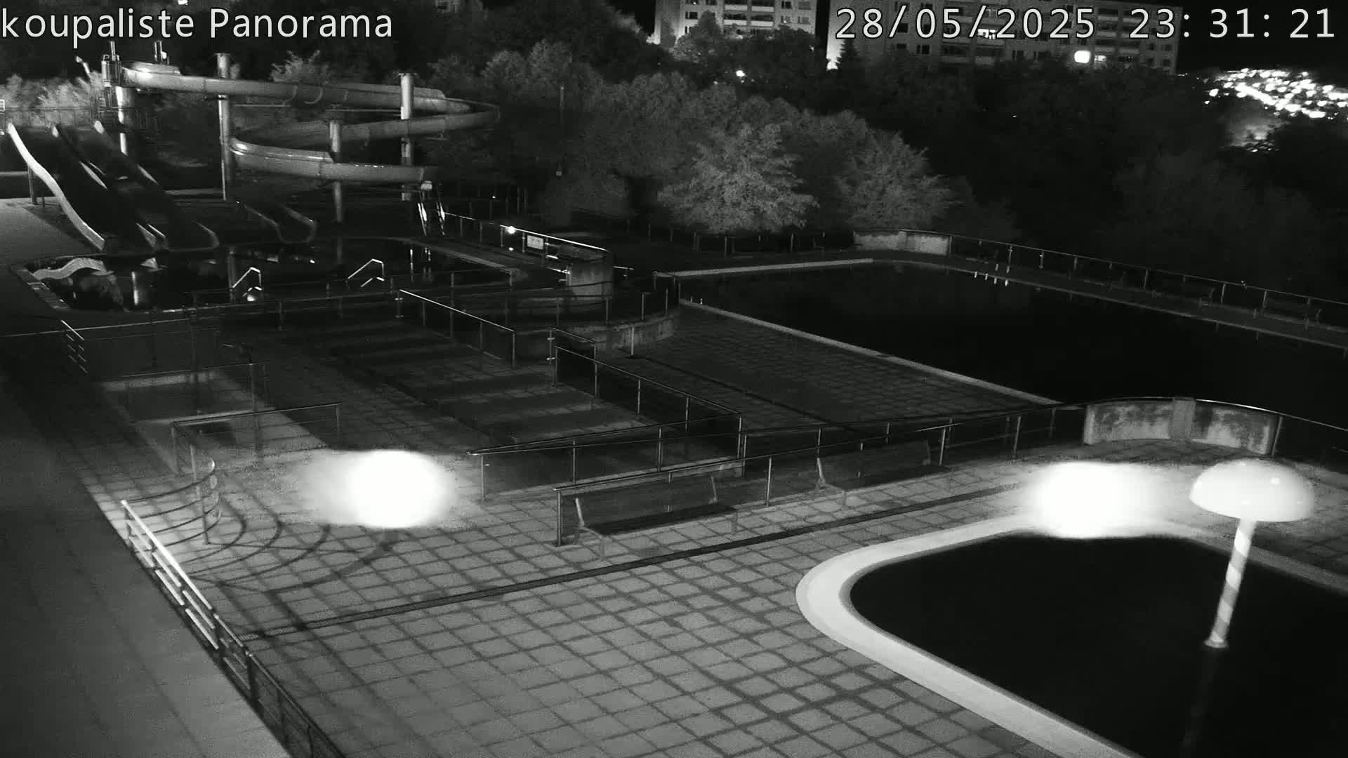 A nighttime, black-and-white overhead view of an empty swimming pool complex with water slides and surrounding trees under dark conditions.