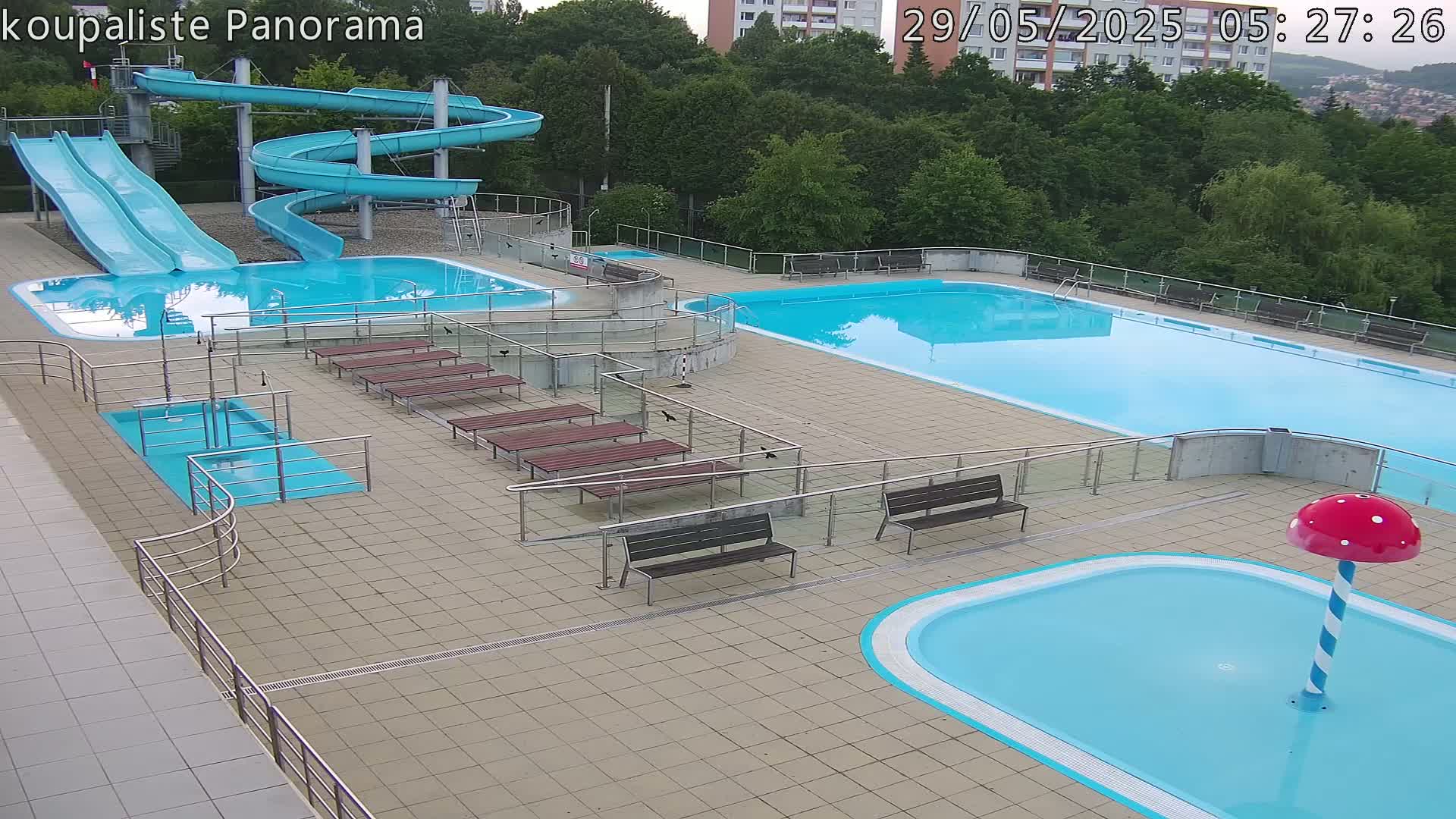An outdoor pool complex with two large pools, several water slides, and various benches, under a partly cloudy sky.