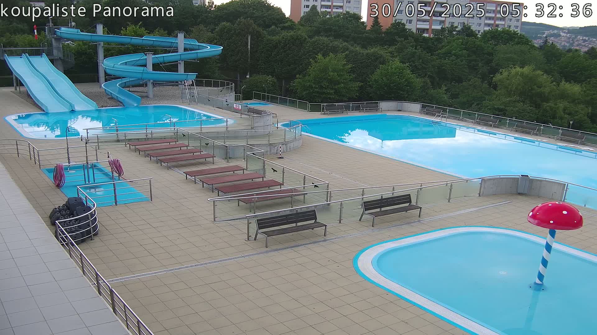 An outdoor pool complex with two large pools, two slides, a small children's pool with a mushroom-shaped water feature, and several benches, all on a tiled deck under a partly cloudy sky.