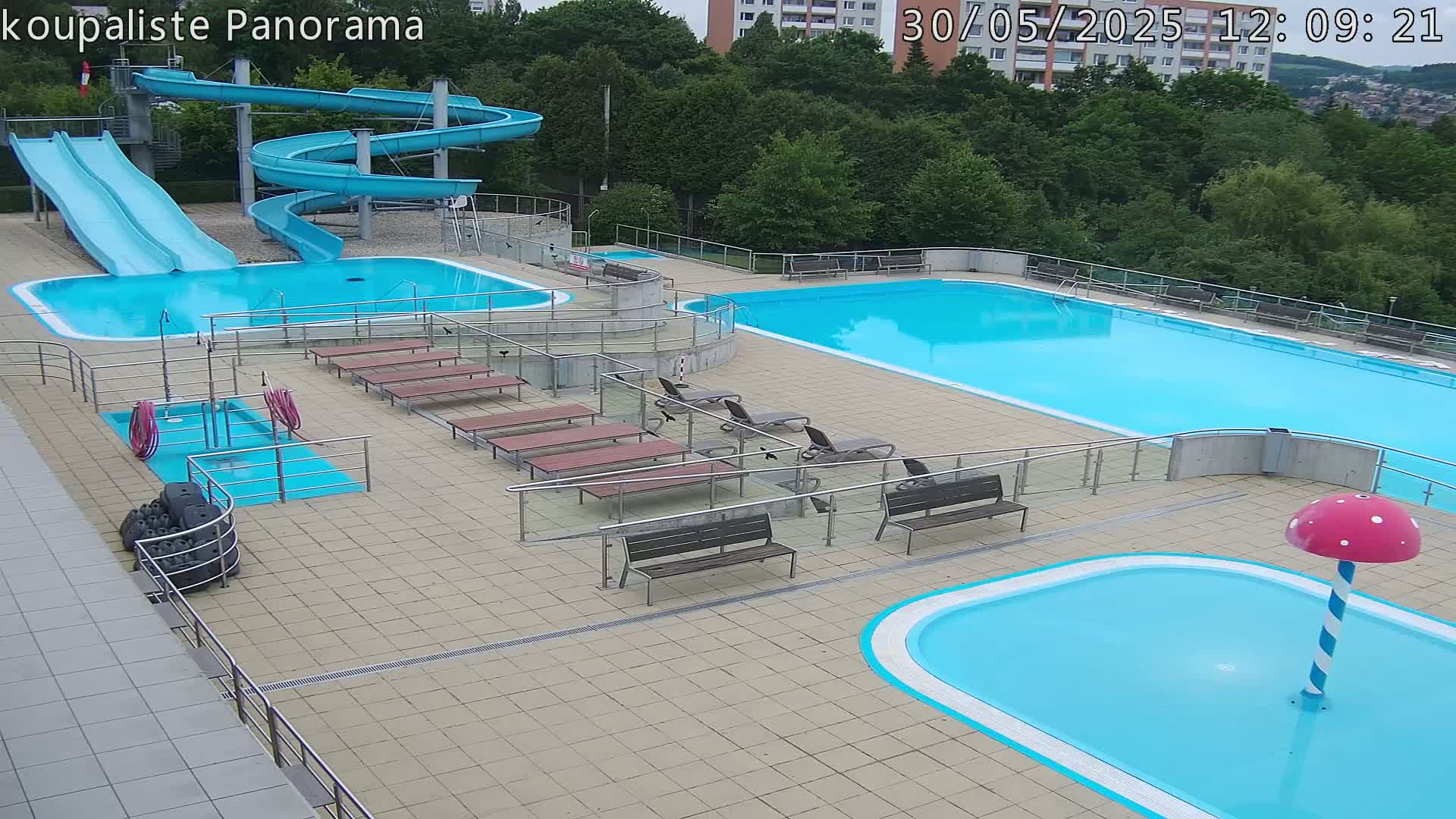 An outdoor swimming complex with several pools, water slides, benches, and a small mushroom-shaped water feature on a mostly sunny day.