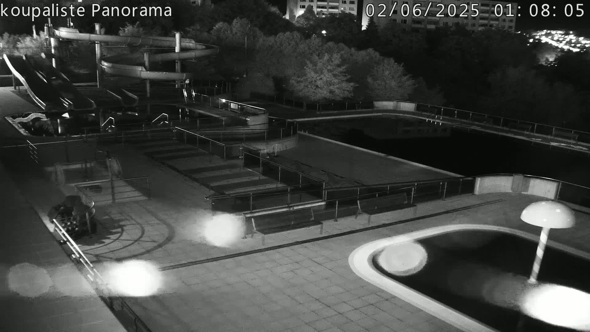 An outdoor nighttime view of an empty water park showing slides, pools, and benches under bright lights.