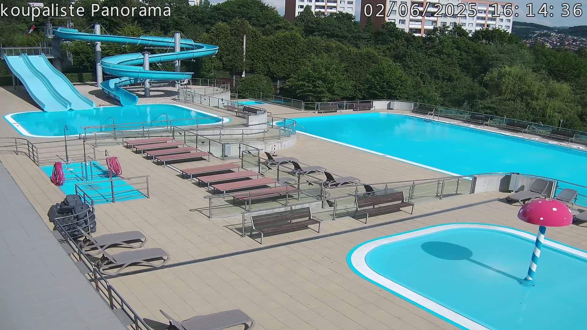 An outdoor pool complex on a sunny day features two large swimming pools, a pair of slides, a small children's pool with a mushroom-shaped water feature, and several lounge chairs and benches.