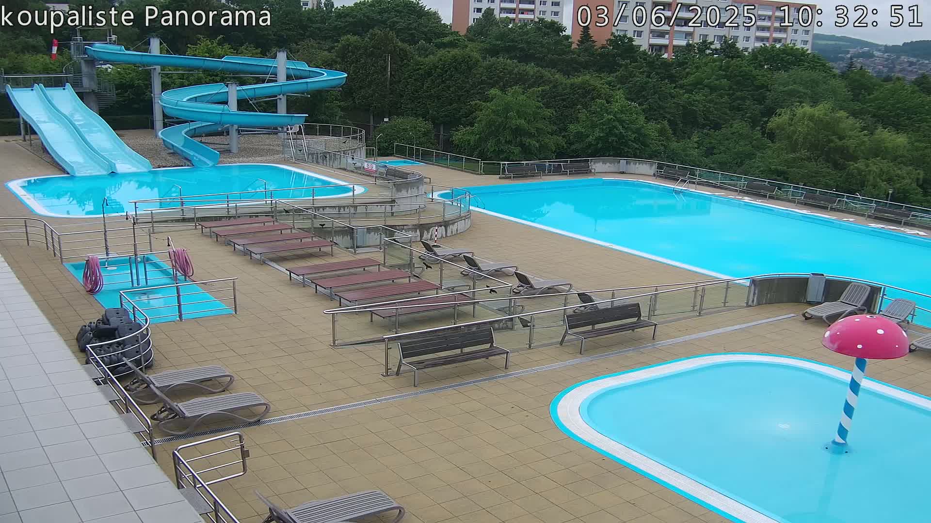 An outdoor swimming pool complex with multiple pools, slides, and lounge chairs on a partly cloudy day.