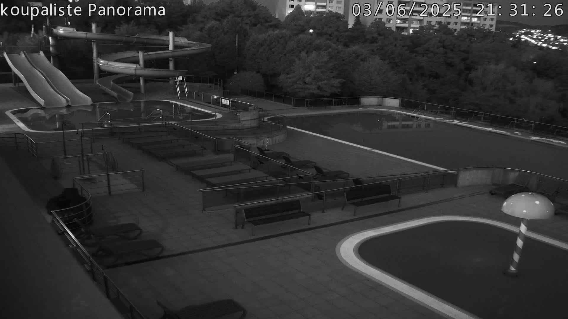 An outdoor nighttime black-and-white view of an empty swimming pool complex with water slides and lounge chairs.
