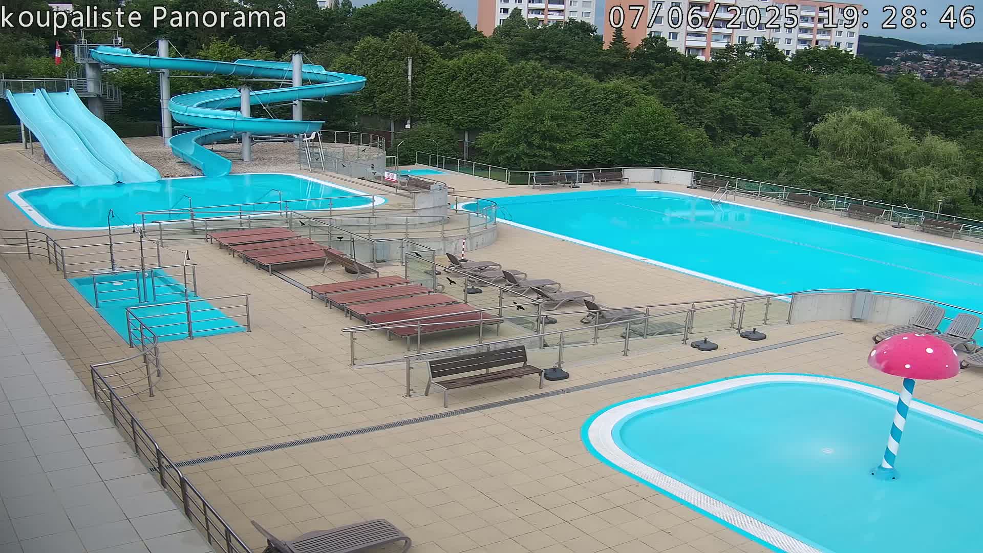 An outdoor pool complex with two large pools, a small children's pool with a mushroom-shaped fountain, two water slides, and several lounge chairs under a partly cloudy sky.