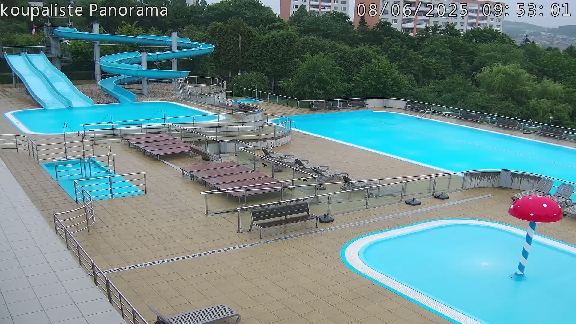 An outdoor pool complex with multiple pools, water slides, and lounge chairs, under an overcast sky.