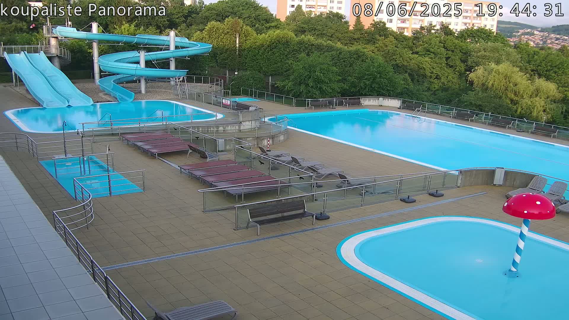 An outdoor pool complex with two large swimming pools, a smaller children's pool with a mushroom-shaped water feature, two water slides, and multiple lounge chairs under a partly cloudy evening sky.