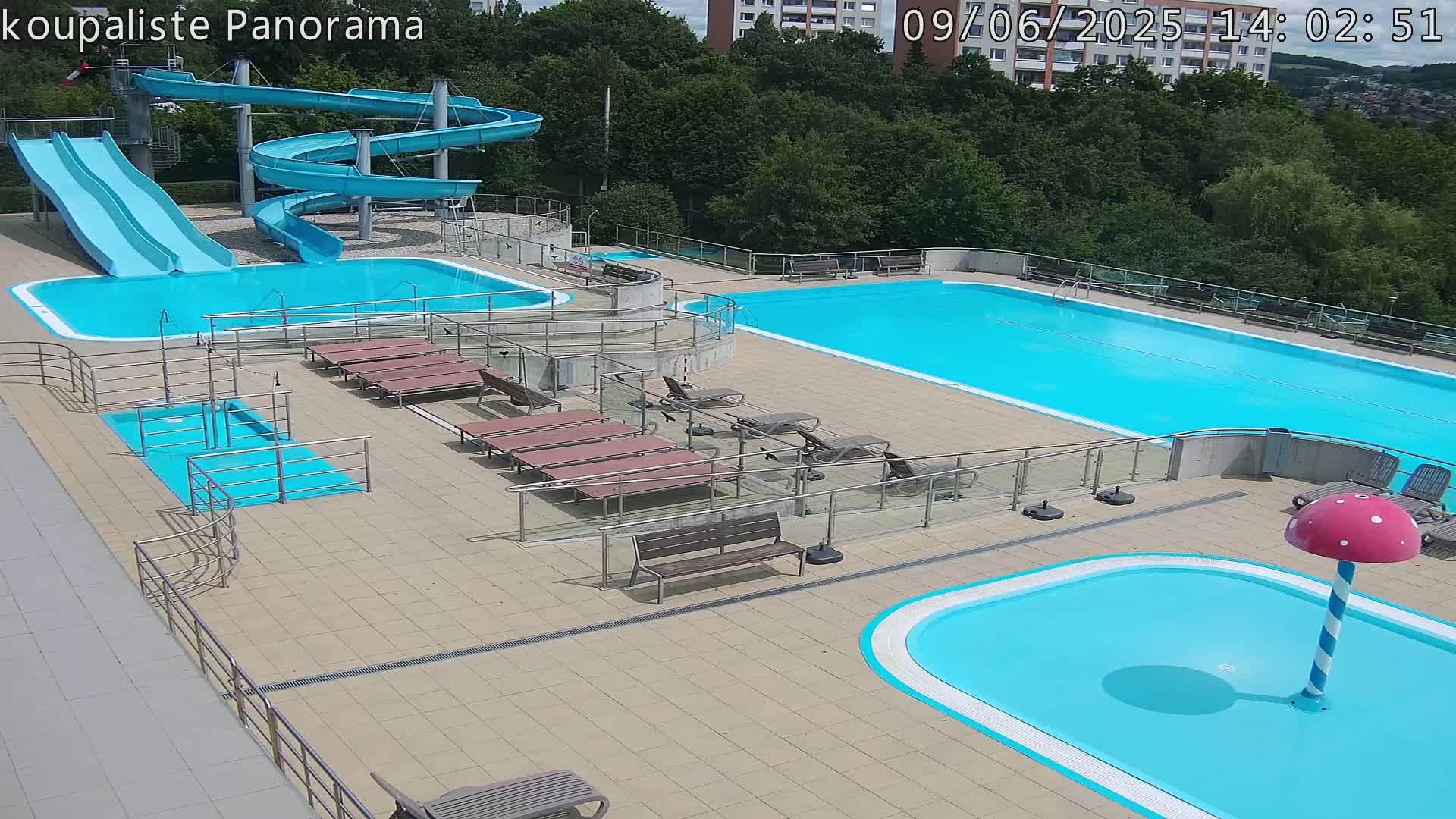 An outdoor pool complex with multiple pools, including a large lap pool, a smaller pool with a mushroom-shaped water feature, and a children's area, features two water slides and numerous lounge chairs on a sunny day.
