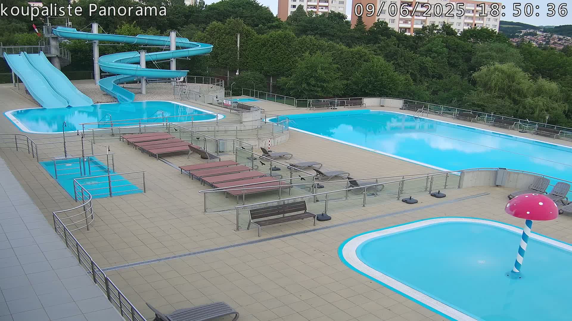 An outdoor pool complex with two large pools, several water slides, lounge chairs, and a small children's pool with a mushroom-shaped water feature, under a partly cloudy sky.