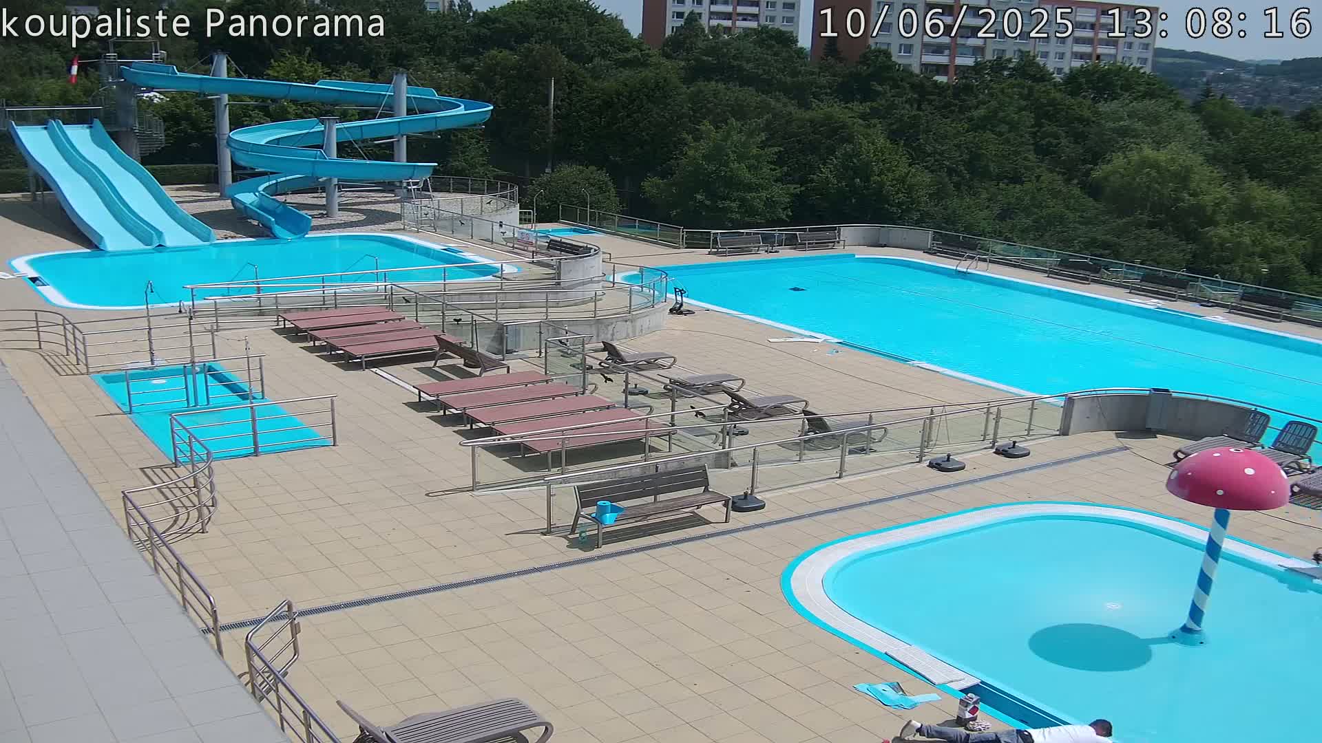 An outdoor pool complex on a sunny day features multiple pools, water slides, lounge chairs, and a mushroom-shaped water feature.
