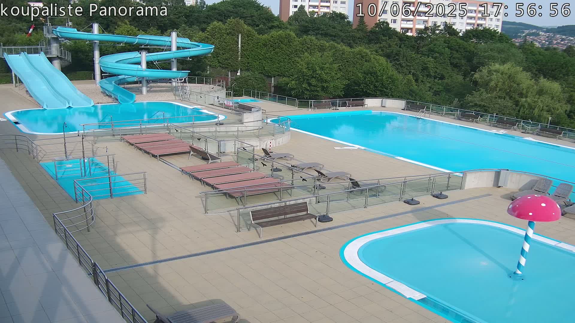 An outdoor pool complex with two large swimming pools, a smaller children's pool with a mushroom-shaped fountain, two water slides, and several lounge chairs under a partly sunny sky.