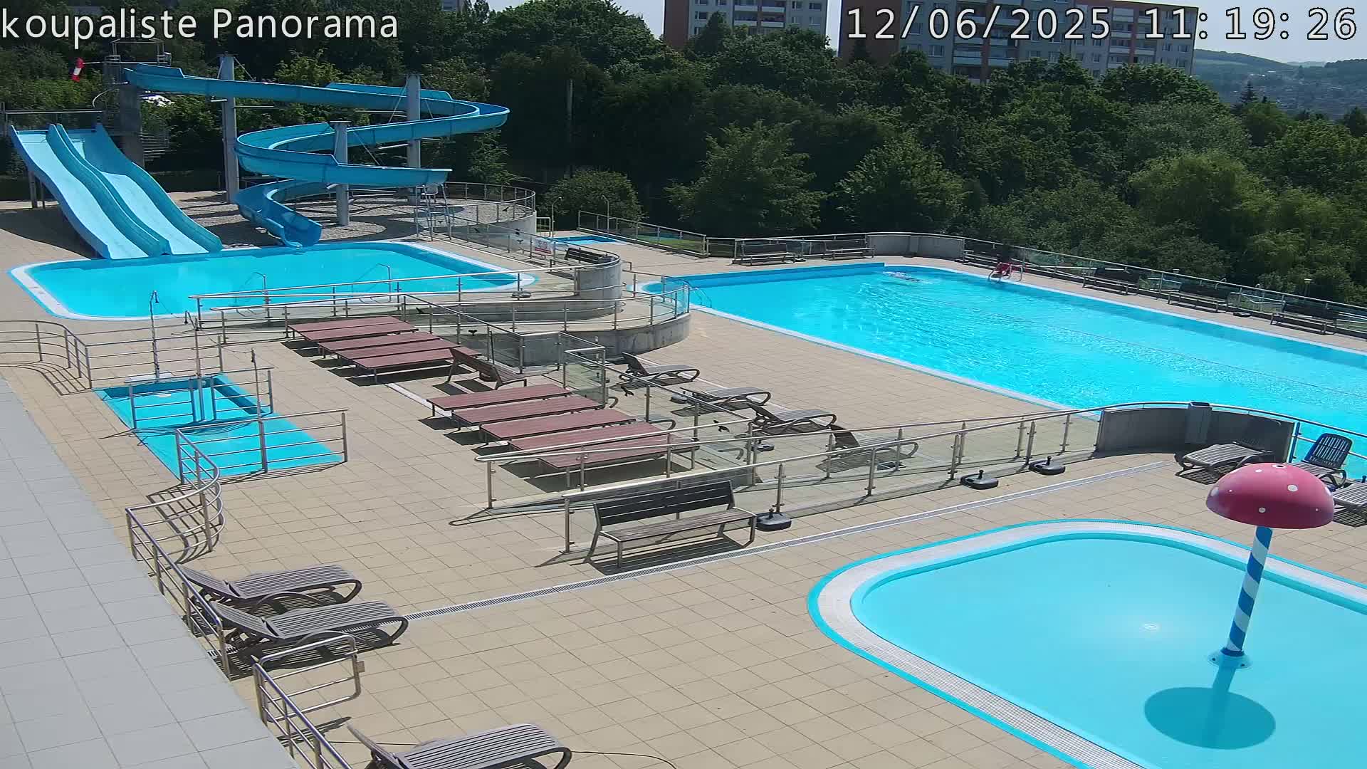 An outdoor pool complex with multiple pools, including a large main pool, smaller pools, and water slides, on a sunny day.