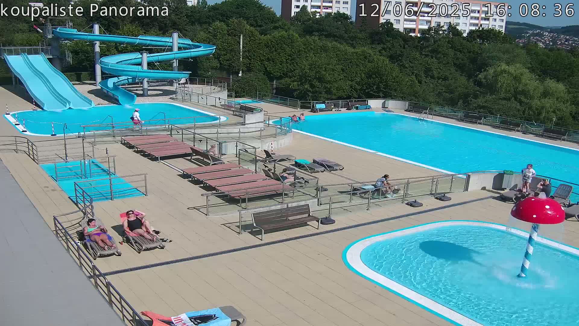 An outdoor pool complex on a sunny day features a large swimming pool, several smaller pools, water slides, and lounge chairs surrounding the pools.
