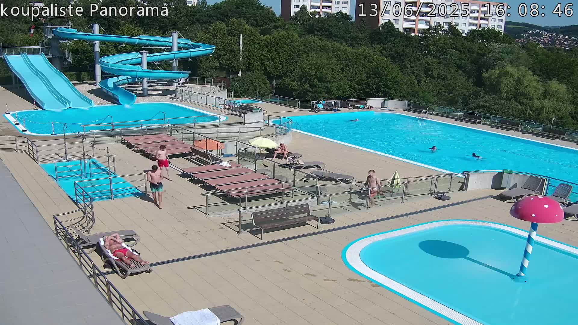 An outdoor swimming pool complex on a sunny day features multiple pools, water slides, lounge chairs, and several people enjoying the facilities.